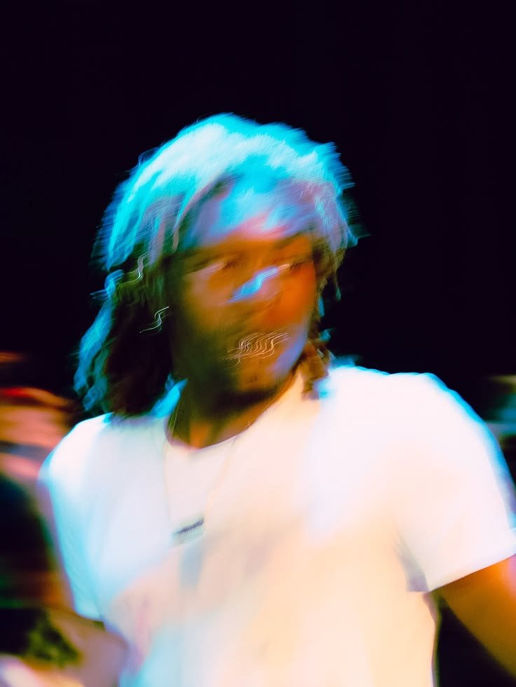 Blurred motion photo of a person with long hair lit in blue stage light, wearing a white t-shirt and layered necklaces.